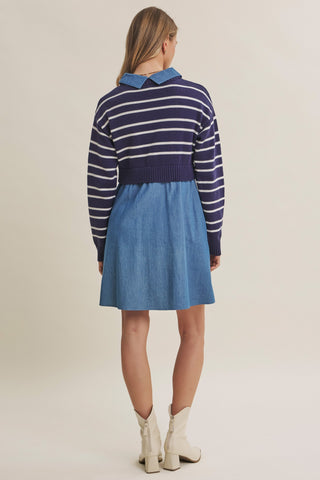 A woman is shown from the back wearing a knit-and-denim dress with a navy and white striped yoke, a blue collar, and a blue denim skirt, paired with off-white ankle boots against a plain beige studio background.