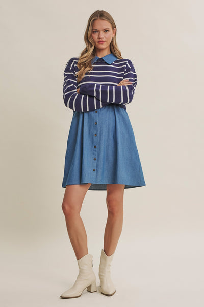 A woman modeling a long-sleeve button-front shirt dress with a navy and white striped yoke and a blue denim skirt, paired with white ankle boots, against a plain beige studio background.