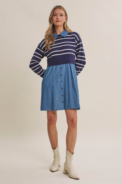 A woman models a long-sleeve knit polo dress with a navy and white striped yoke and a blue denim button-front skirt, paired with white ankle boots, against a solid light beige background.