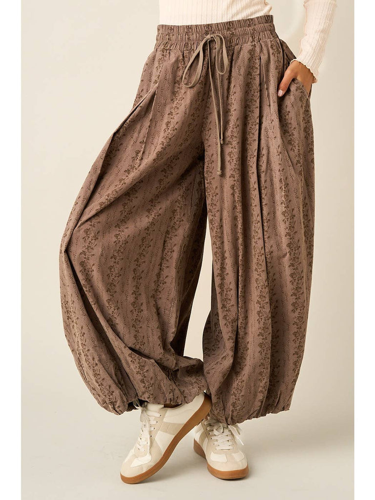 A person wearing loose, balloon-style brown pants with a subtle floral print, an elastic drawstring waist, and side pockets, paired with white sneakers against a plain light beige studio background.