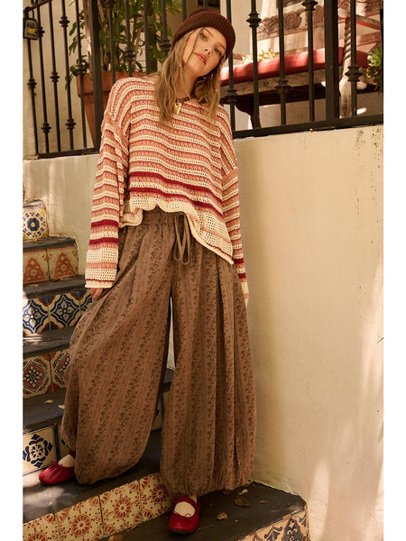 A woman stands on outdoor tiled steps wearing a cream and rust striped knit sweater, brown wide-leg patterned pants, a brown beanie, and red shoes.