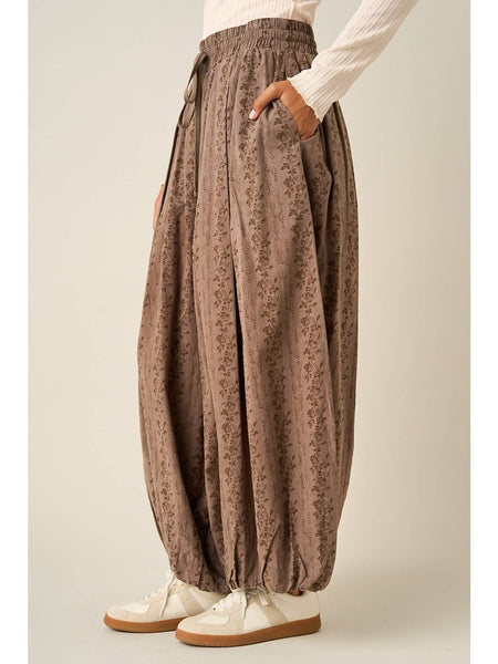 A person wearing loose, high-waisted brown patterned harem-style pants with an elastic waist and drawstring, paired with a cream ribbed long-sleeve top and white sneakers, against a plain light beige studio background.
