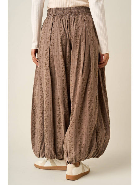 A person wearing loose, pleated harem-style pants in a taupe-brown color with a subtle floral pattern, paired with a white ribbed long-sleeve top and white slip-on shoes, shown from the waist down against a plain light beige studio background.