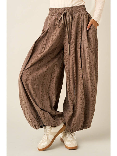 A person wearing loose, balloon-style brown pants with a subtle floral print, an elastic drawstring waist, and side pockets, paired with white sneakers against a plain light beige studio background.