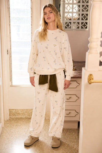 Woman wearing a cream-colored outfit with a pattern, standing in a room with a window and furniture.