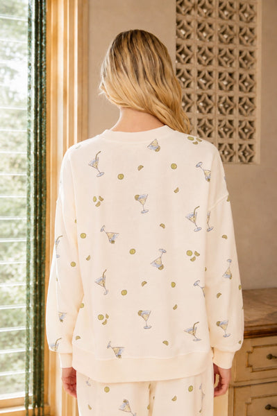 Woman wearing a cream-colored pajama set with cocktail-themed pattern in a room with a window and decorative wall.