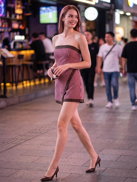 A woman walking on a city street at night wearing a strapless mini dress with vertical pink and white stripes and dark brown trim, paired with dark brown high heels.