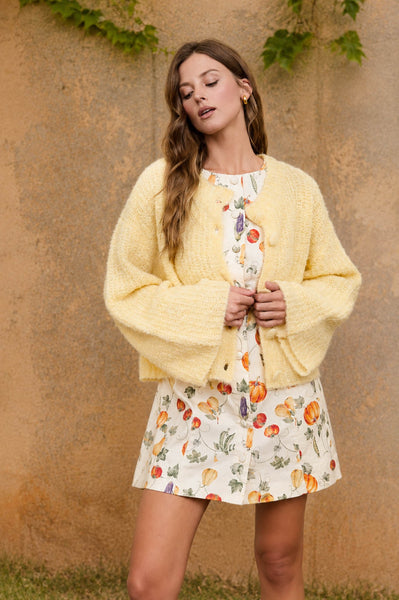 A woman stands outdoors against a textured tan wall wearing a pale yellow fuzzy knit cardigan over a white dress with a colorful fruit print and a matching short skirt.