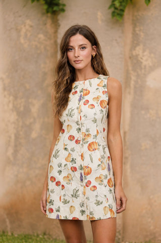 A woman modeling a sleeveless A-line dress with a white base and a colorful fruit print, standing against a textured beige wall.