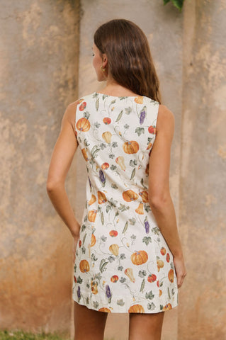 A woman viewed from behind wearing a sleeveless white dress with an all-over autumn harvest print of pumpkins, gourds, and vegetables, set against a rustic textured stone wall.