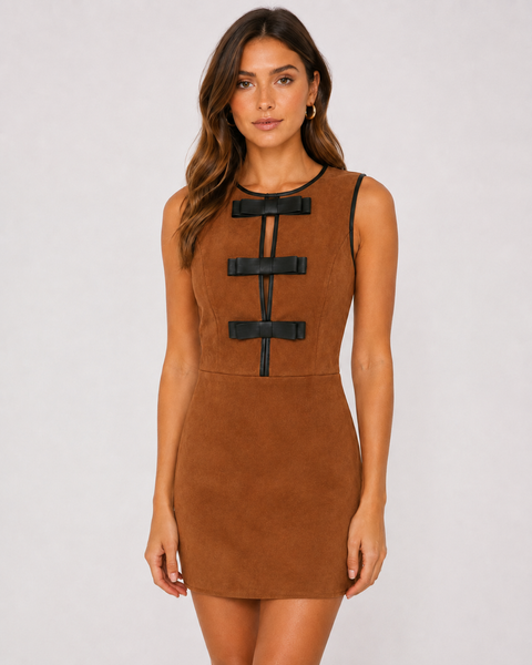 A woman modeling a sleeveless brown faux-suede mini dress with black strap details down the front, photographed against a light gray studio background.