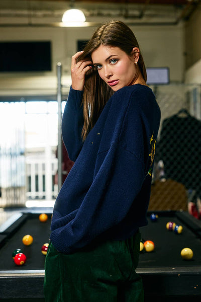 Woman in a blue sweater standing in front of a pool table with pool balls.