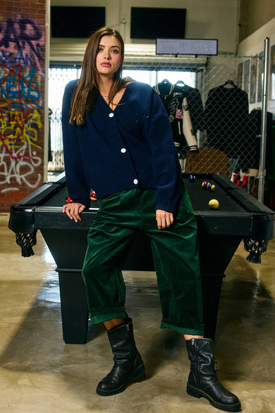 Person wearing a navy cardigan and green pants sitting on a pool table in an indoor setting with graffiti on the wall.