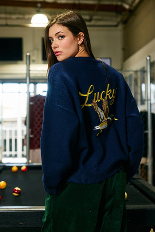 Woman wearing a blue sweater with 'Lucky' and an eagle design, standing in front of a pool table.