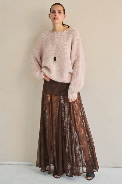 A woman stands against a plain beige wall wearing a fuzzy light pink sweater and a long brown sheer lace skirt.