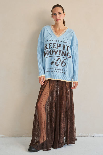 Woman wearing a light blue sweater with text and a brown lace skirt against a beige background