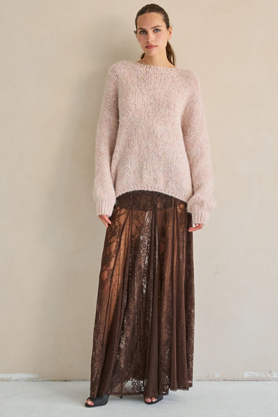 A woman stands against a plain beige wall wearing a fuzzy light pink knit sweater and a long brown pleated skirt with a sheer floral overlay.