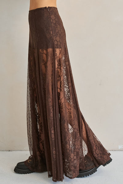 A brown lace maxi skirt with sheer mesh side panels, shown on a model from the waist down wearing black platform boots against a plain beige wall.