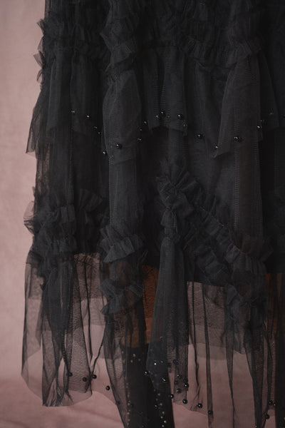 A close-up of a black sheer tulle dress with layered ruffles and small black bead embellishments, shown against a dusty mauve-pink background.