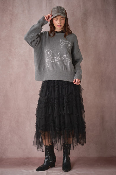 A model wearing a gray knit sweater with white graphic text, a long black tiered tulle skirt, and black ankle boots, standing against a textured mauve-brown studio backdrop.