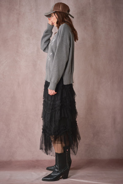 Woman wearing a gray sweater, black tulle skirt, and black boots against a plain background