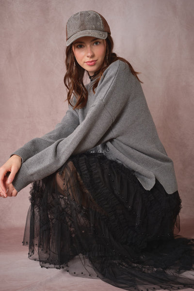 A woman wearing a gray knit sweater, black tulle skirt, and gray cap, posing on a dusty mauve studio backdrop.