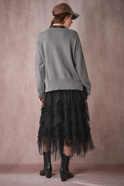 A model shown from the back wearing a gray knit sweater, a long black ruffled tulle midi skirt, sheer black tights, and black lace-up ankle boots, set against a textured dusty pink studio backdrop.