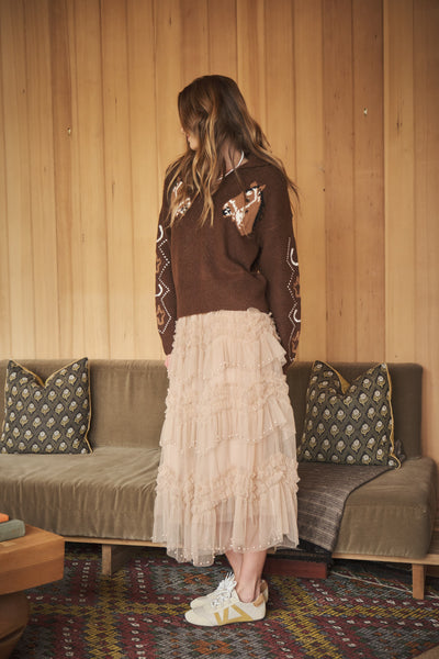 A woman stands with her back to the camera in a cozy room with wood-paneled walls, wearing a brown knit sweater with embroidered details, a long cream ruffled midi skirt, and white sneakers.