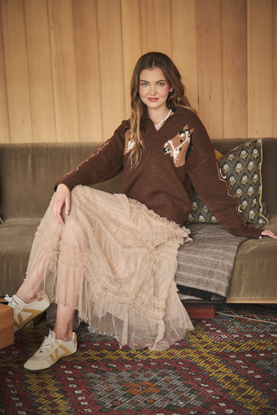 A woman sits on a brown velvet sofa wearing a brown knit sweater with a fox patch, a long beige tulle skirt, and white sneakers with gold stripes, set against a wood-paneled wall.