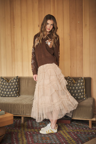 A woman stands in a rustic room with wood-paneled walls, wearing a brown knit sweater with white embroidery and a long, tiered beige tulle midi skirt. She is posed in front of a brown sofa with patterned pillows.