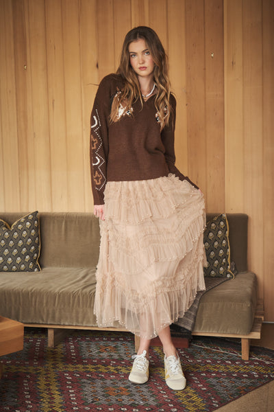 A woman stands in a room with wood-paneled walls, wearing a brown knit sweater with embroidered details on the sleeves and a long, tiered ruffled midi skirt in a light beige or cream color. She is wearing white sneakers and standing in front of a greenish-brown sofa with patterned pillows.