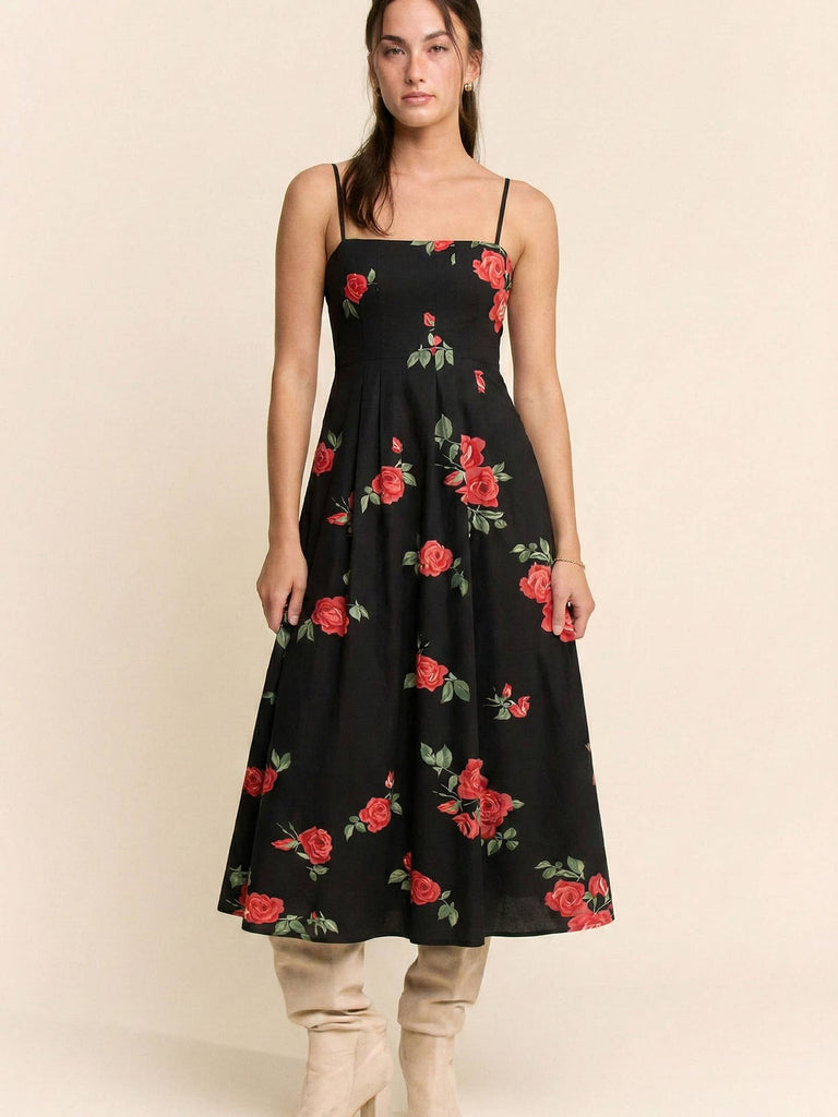 A woman modeling a black midi dress with a red rose and green leaf floral pattern, paired with beige boots, against a solid beige background.
