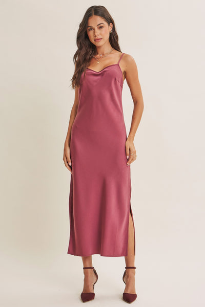 A woman modeling a sleeveless satin midi slip dress in a dusty rose pink color, paired with dark brown strappy heels, against a plain light beige studio background.