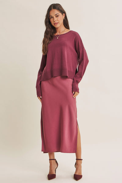 A woman modeling a matching two-piece outfit consisting of a long-sleeve crewneck sweater and a high-slit midi skirt in a dusty rose or mauve shade, paired with matching pointed-toe heels against a plain light beige studio background.