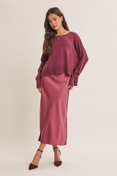 A woman modeling a dusty rose two-piece outfit consisting of a long-sleeve cropped knit top and a matching midi skirt, paired with dark brown pointed-toe heels against a plain light beige studio background.
