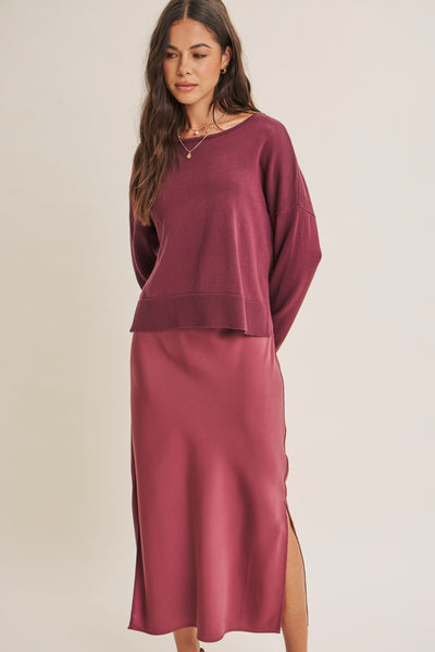 A woman modeling a matching two-piece outfit consisting of a long-sleeve crewneck top and a high-slit midi skirt in a dusty rose or mauve color, set against a plain light beige studio background.