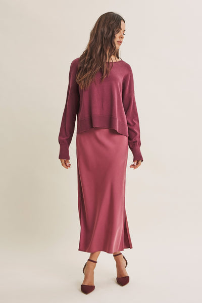 A woman modeling a two-piece dusty rose knit set consisting of a long-sleeve cropped sweater and a matching midi skirt, paired with dark brown pointed-toe heels against a plain light beige studio background.