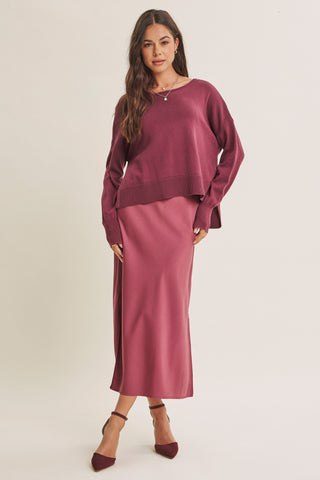 A woman modeling a matching two-piece outfit consisting of a long-sleeve crewneck top and a midi skirt in a dusty rose or mauve shade, paired with dark brown pointed-toe heels against a plain light beige studio background.