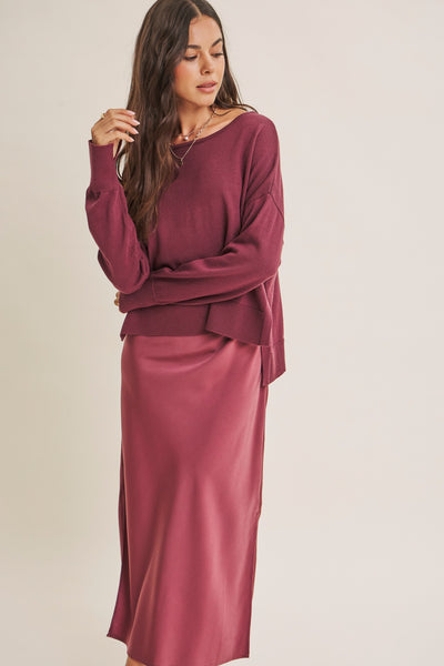 A woman modeling a matching two-piece outfit consisting of a long-sleeve knit top and a satin midi skirt in a dusty rose or mauve color, set against a plain light beige studio background.