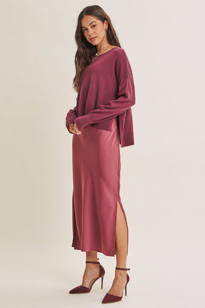 A woman modeling a long-sleeve midi dress in a dusty rose or mauve color, paired with matching pointed-toe heels, against a plain light beige studio background.