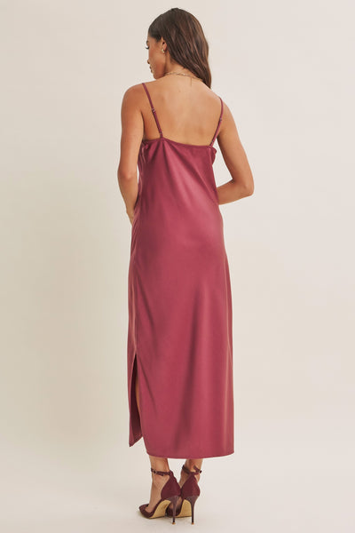 A woman shown from the back wearing a satin slip dress in a dusty rose color, paired with dark brown strappy heels, against a plain light beige studio background.