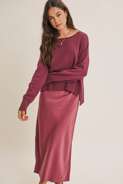 A woman modeling a matching two-piece outfit consisting of a long-sleeve crewneck top and a satin midi skirt in a dusty rose or mauve shade, set against a plain light beige studio background.
