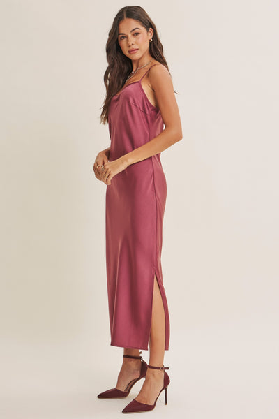 A woman modeling a satin slip dress in a dusty rose color, paired with matching burgundy high heels, against a plain beige studio background.