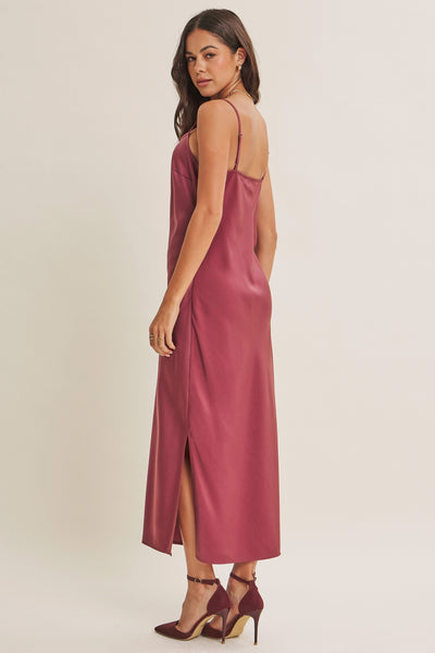 A woman modeling a dusty rose satin slip dress with spaghetti straps and a high side slit, paired with matching heels, against a plain beige studio background.
