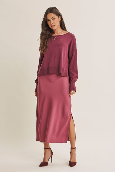 A woman modeling a matching two-piece outfit consisting of a long-sleeve crewneck top and a high-slit midi skirt in a deep berry or mauve shade, paired with matching pointed-toe heels against a plain light beige studio background.
