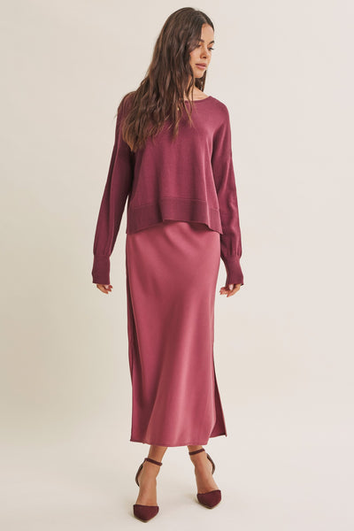 A woman modeling a matching two-piece knit set consisting of a long-sleeve cropped sweater and a high-waisted midi skirt in a dusty rose or mauve shade, paired with dark brown pointed-toe heels against a plain light beige studio background.
