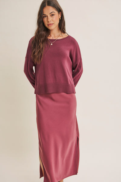 A woman modeling a dusty rose two-piece knit set consisting of a cropped long-sleeve top and a matching midi skirt with side slits, set against a plain light beige background.
