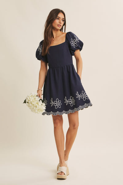 A woman modeling a navy blue mini dress with puff sleeves, white floral embroidery, and a scalloped lace hem, holding a bouquet of white flowers against a solid light beige background.
