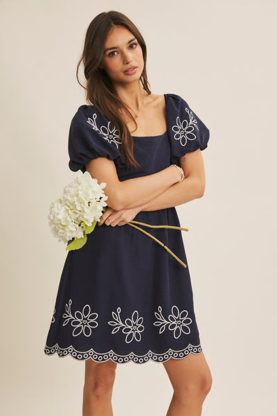 A woman modeling a navy blue dress with white floral embroidery and puff sleeves, holding a bouquet of white flowers against a plain beige studio background.