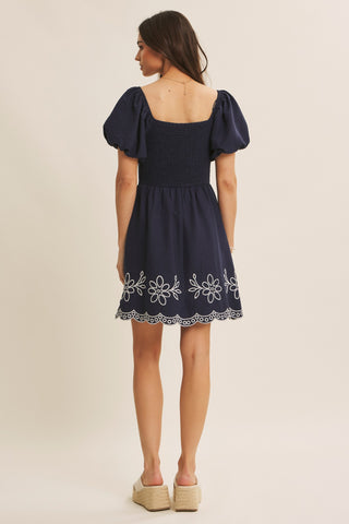 A woman modeling a navy blue puff-sleeve mini dress from the back, featuring white floral embroidery along the hem, set against a plain beige studio background.